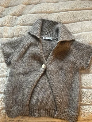  stickad kofta från zara - Mysig kortärmad stickad kofta i melerad brun färg från zara. Koftan har en stor krage, en knapp framtill och ribbade detaljer vid ärmslut och nederkant.