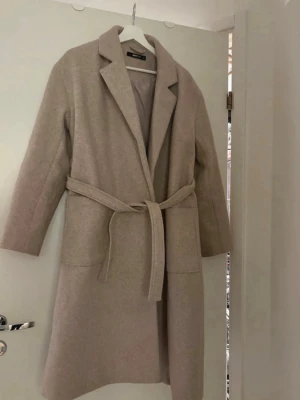 Beige kappa från Gina Tricot - Stilren beige kappa från Gina Tricot med klassisk krage och knytskärp i midjan. Kappan har raka ärmar, två fickor framtill och är helfodrad för extra komfort. Perfekt för dig som vill ha en enkel men snygg look till hösten eller våren. Fläck på baksida 