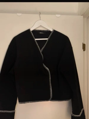 Svart cardiganjacka med kontrastsömmar - Stilren svart cardiganjacka från Gina Tricot med vita kontrastsömmar längs kanterna. Jackan har rak passform, lång ärm och öppen front utan knappar. Perfekt för lager på lager och enkel att matcha med olika outfits.