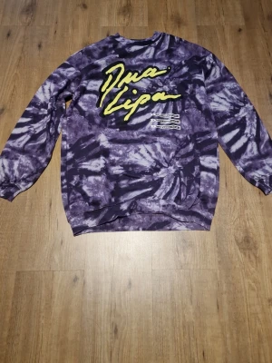 Lila Dua Lipa sweatshirt - Cool lila och svart tie-dye sweatshirt med Dua Lipa-logga i gult framtill. Tröjan har rund hals, långa ärmar och ribbade muddar. Perfekt för dig som gillar streetstyle och vill sticka ut med en unik design.
