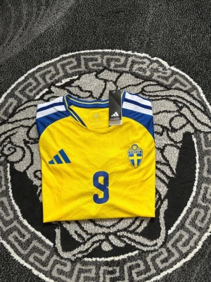 Svensk landslagströja Adidas gul - Gul svensk landslagströja från Adidas med blå detaljer och nummer 9 på bröstet. Tröjan har korta ärmar, blå och vita ränder på axlarna samt det svenska fotbollsförbundets märke på vänster sida. Tillverkad i lätt och ventilerande material.