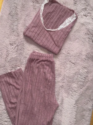 Rosa ribbat loungewear-set med spets - Mysigt loungewear-set i mjukt ribbat material i dammigt rosa. Setet består av ett linne med vita spetsdetaljer vid halsen och matchande byxor med resår i midjan. Perfekt för chill dagar hemma.