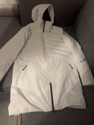 Vit dunjacka från Snö of Sweden - Snygg vit dunjacka från Snö of Sweden med huva och dragkedja framtill. Jackan har två sidofickor med dragkedja och är tillverkad i GORE-TEX-material som står emot både vind och vatten. Perfekt för kalla vinterdagar.