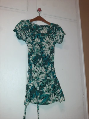 Blommig kortklänning med knyt - Kortklänning med stort blommönster i vitt, turkos och grönt. Klänningen har korta puffärmar, rund halsringning och ett knytband i midjan för en snygg passform. Perfekt för dig som gillar färg och mönster. Storlek M/L