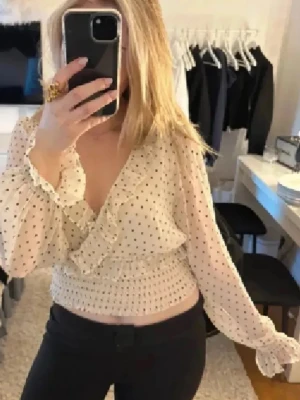 Prickig volangblus från Bik Bok - Söt vit blus med svarta prickar från Bik Bok. Blusen har djup v-ringning med volangdetaljer, smockad midja och långa, transparenta ärmar. Perfekt för dig som gillar romantisk och trendig stil. Materialet är lätt och luftigt.