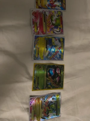 Pokémonkort samling holografiska - Fem stycken Pokémonkort med holografisk yta i olika färger och motiv. Korten har färgstarka illustrationer och detaljerade mönster, flera med guld- och regnbågseffekter. Perfekt för samlare eller Pokémonfans som vill utöka sin kortsamling.