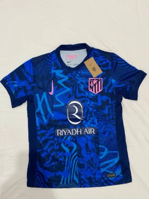 En Atletico Madrid tredjeställ från 24/25 - Priset kan diskuteras. Snygg blå matchtröja från Atlético Madrid med grafiskt mönster i olika blå nyanser, rosa Nike-logga och klubbmärke på bröstet. Sponsorlogga 'Riyadh Air' på framsidan. Tillverkad i lätt och ventilerande material, perfekt för träning eller match.