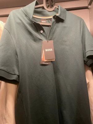Mörkgrön pikétröja från BOSS - Snygg mörkgrön pikétröja från BOSS med klassisk krage och korta ärmar. Diskret BOSS-logga broderad på bröstet och två knappar vid halsen. Perfekt till både jeans och chinos för en clean look. Aldrig använd :)