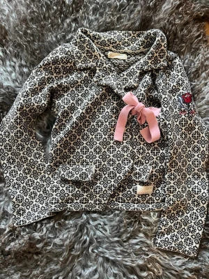 Mönstrad kofta Odd Molly med rosett - Unik svartvit mönstrad kofta från Odd Molly med lång ärm, rosa rosett vid halsen och broderad röd ros på bröstet. Koftan har två fickor framtill och snygg krage. Perfekt för dig som gillar detaljer och sköna material.