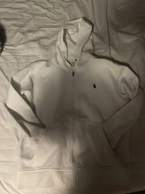 Vit hoodie från Polo Ralph Lauren - Säljer en stilren vit hoodie från Polo Ralph Lauren med dragkedja framtill och klassisk huva. Tröjan har fickor på magen och den ikoniska lilla loggan broderad på bröstet. Perfekt för dig som gillar enkel och clean streetwear. Storlek S men passar även M