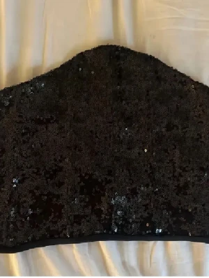 Svart paljett bandeau tubtopp - Säljer en svart bandeau tubtopp täckt med glittriga paljetter. Toppen har snörning i ryggen för justerbar passform och är perfekt för att sticka ut. Materialet känns syntetiskt och toppen är axelbandslös.