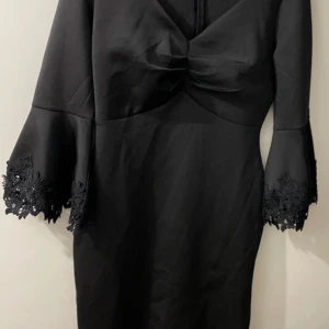 Svart klänning med spetsärmar - Elegant svart klänning med v-ringning och trumpetärmar dekorerade med spetsdetaljer. Figurnära passform och knälång modell som ger en stilren siluett. 