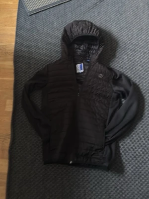 Svart vindjacka från Jack & Jones - Snygg svart vindjacka från Jack & Jones med huva och dragkedja. Jackan har quiltade partier framtill och på huvan, samt släta ärmar. Perfekt för blåsiga dagar och enkel att matcha med olika outfits.