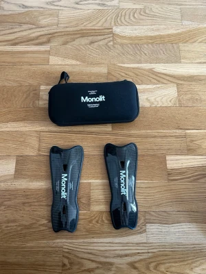 Monolit benskydd i svart kolfiber - Säljer ett par Monolit benskydd i svart kolfiber med vit logga. De har en ergonomisk form och kommer med ett skyddande fodral. Perfekta för dig som vill ha lätta och slitstarka benskydd till fotbollsträningen.