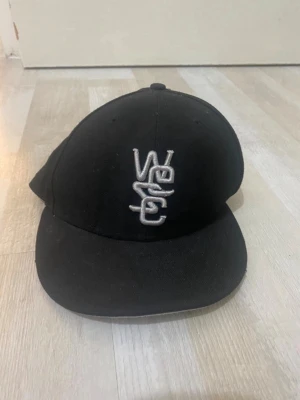 Svart keps med broderad logga - Säljer en svart keps från WeSC New Era med böjd skärm och broderad vit logga framtill. Klassisk modell med ventilation på ovansidan och diskret svart logga på sidan. Tillverkad i slitstarkt tyg, perfekt för en streetwear-look.