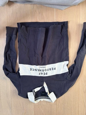 Svart långärmad Peak Performance  - Svart långärmad henley-skjorta från Peak Performance med vit krage och bred vit rand över bröstet med logga. Skjortan har knappar vid halsen och är gjord i mjuk bomull för en chill och sportig vibe.