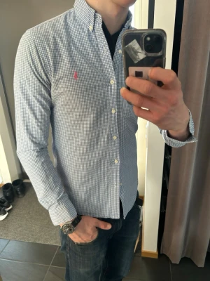 Blårutig skjorta från Ralph Lauren - Klassisk blårutig skjorta från Ralph Lauren med button-down krage och lång ärm. Skjortan har små vita och blå rutor samt en rosa broderad logga på bröstet. Tillverkad i bomull och stängs med vita knappar framtill.