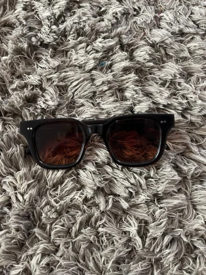 Svarta solglasögon med fyrkantig form - Snygga svarta solglasögon från CHIMI med bred, fyrkantig båge och mörka glas. Klassisk design som passar till det mesta och ger en cool vibe. Bågarna har små silverdetaljer vid sidorna för extra stil.