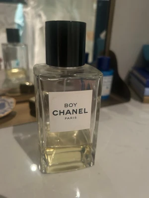 Boy Chanel parfym 200ml för byte. 40/200ml kvar - Boy Chanel parfym i en stilren, rektangulär glasflaska med svart plastlock. Flaskan är transparent och visar den ljusgula vätskan inuti. Märkets klassiska etikett framtill ger en lyxig känsla och designen är minimalistisk och elegant.