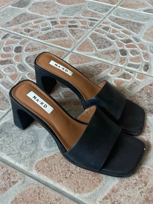 Svarta mules från NA-KD - Stilrena svarta mules från NA-KD med bred rem och fyrkantig tå. Skorna har en chunky klack och innersula i brunt konstläder. Perfekta till allt. Endast använda vid ett tillfälle 🌻🤝