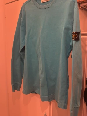 Blå långärmad tröja Stone Island - Snygg blå långärmad tröja från Stone Island med klassisk patch på ärmen. Tröjan har rund halsringning och är gjord i mjukt bomullsmaterial. Perfekt för dig som gillar streetwear och vill ha en clean look.