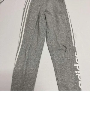 Gråa mjukisbyxor från Adidas - Snygga gråa mjukisbyxor från Adidas med klassiska vita ränder längs benen och stor Adidas-logga på ena benet. Tillverkade i mjukt och bekvämt material, perfekta för chill eller träning. Normal passform och resår i midjan.