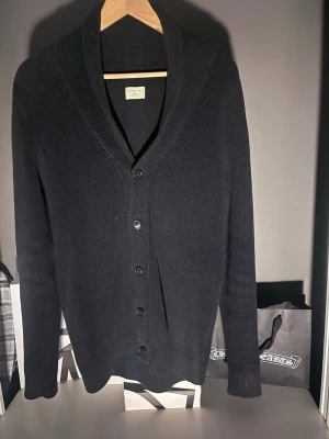 Svart ribbstickad kofta - Stilren svart ribbstickad kofta från selected homme med sjalkrage och knäppning framtill. Koftan har långa ärmar och en avslappnad passform, perfekt att slänga över en t-shirt. Materialet är mjukt och håller värmen bra.