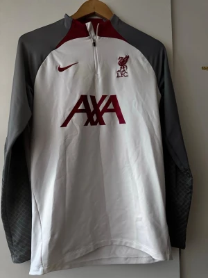 Liverpool Nike träningströja vit/grå - Snygg långärmad träningströja från Nike för Liverpool FC. Tröjan är vit med grå ärmar och vinröda detaljer, halv dragkedja framtill och broderad LFC-logga på bröstet. AXA sponsortryck i vinrött. Perfekt för fotbollsträning eller sportig stil.