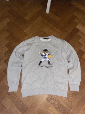 Polo bear Ralph Lauren  - Grå sweatshirt från Polo Ralph Lauren med klassisk rund hals och ribbade muddar. På bröstet finns en tryckt Polo Bear i basebolloutfit. Mjuk bomullsblandning som känns skön mot huden. Perfekt för en avslappnad och trendig stil.