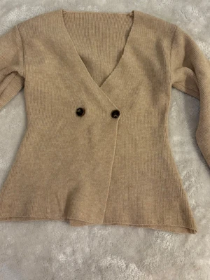 Beige stickad omlottkofta - Säljer en beige stickad omlottkofta med två mörka knappar framtill. Koftan har djup V-ringning och långa ärmar. Materialet är mjukt och ribbstickat, perfekt för lager på lager. Enkel och stilren design utan synligt märke.