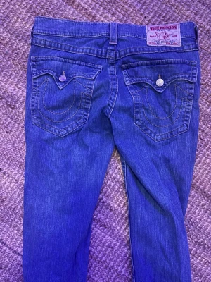 Blå True Religion jeans med kontrastsömmar - Säljer ett par klassiska blå jeans från True Religion med ikoniska kontrastsömmar på bakfickorna och snygga detaljer. Jeansen har fem fickor, bälteshällor och knappgylf.  Dom är sönder på botten men går att ha på någon som är 175 hade kunnat ha dom och säljer dom för de är för stora för mig