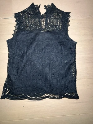 Marinblå  spetsblus med hög hals - Snygg marinblå  blus i spets med hög hals och detaljerat mönster. Blusen är ärmlös och har en elegant, genombruten design som ger en trendig look. Perfekt att styla med jeans eller kjol för en trendig fin outfit💓🍾