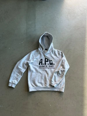APC hoodie - Snygg grå hoodie från A.P.C. med svart tryck på bröstet där det står 'A.P.C. SINCE 1987'. Klassisk passform med huva och ribbade muddar vid ärmar och nederkant. Perfekt för en avslappnad och stilren look. Skriv vid frågor