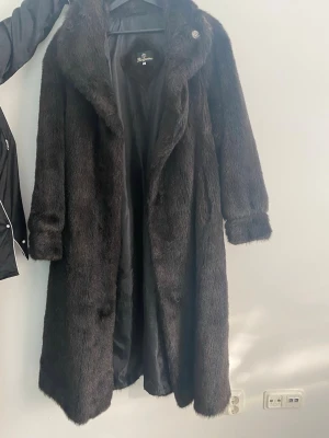 Lång brun pälsjacka med krage - Säljer en lång, brun pälsjacka. Jackan har bred krage, helbrunt foder och stängs med knappar framtill. Klassisk rak modell med långa ärmar och mjuk, glansig päls som ger en lyxig känsla. Perfekt för kalla vinterdagar.