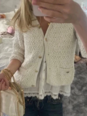 Krämvit kofta från Zara - Säljer en krämvit kofta från Zara med v-ringning och dekorativa guldknappar. Koftan har ett grovt stickat mönster, två små fickor framtill och trekvartsärmar. Perfekt att slänga över en topp för en chic look.