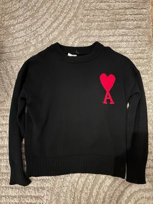Svart stickad tröja från Ami Paris - Snygg svart stickad tröja från Ami Paris med röd hjärtlogga och bokstaven A på bröstet. Tröjan har rund halsringning, ribbade muddar och är gjord i mjukt material som passar perfekt till höst och vinter.