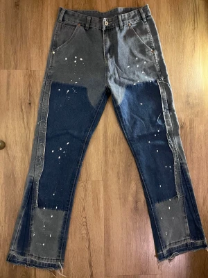 Patchwork jeans med färgstänk - Unika patchwork jeans i grått och mörkblått med råa kanter och vita färgstänk. Byxorna har raka ben, flera fickor och en cool kontrast mellan de olika denimtygerna. Perfekta för dig som gillar streetwear och vill 