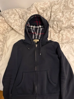 Mörkblå hoodie från Burberry - Säljer en mörkblå hoodie från Burberry med dragkedja och klassiskt rutigt foder i huvan. Hoodien har fickor framtill och justerbar dragsko i huvan. Perfekt för dig som gillar stilrena och exklusiva plagg.