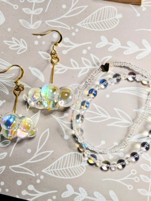 Paket! Moln Örhängen & Armband med genomskinliga regnbågsskimrande pärlor - Paket med örhängen i akryl och Snygga armband med små, genomskinliga pärlor som skimrar i regnbågens färger. Armbandet har ett litet hjärta i metall som detalj. Perfekt accessoar för dig som gillar en lekfull och trendig stil. Högkvalitativ elastisk tråd och 18 cm långt.    Örhängena är fästa på guldpläterade krokar.