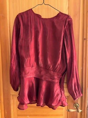Vinröd blus med volang från Gina Tricot - Supersnygg vinröd blus från Gina Tricot med långärmad design och bred midjedel. Blusen har volangdetaljer nedtill och är gjord i ett glansigt, lätt material som ger ett elegant intryck. Perfekt att styla med jeans eller kjol.