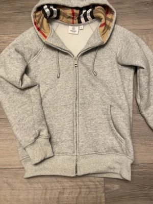 Grå zip-hoodie från Burberry - Snygg grå hoodie från Burberry med dragkedja och klassiskt rutigt foder i huvan. Mjukt material, ribbade muddar och fickor framtill. Perfekt för en stilren och avslappnad look.