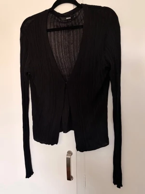 Svart ribbad kofta/tröja - Tunn svart ribbad kofta från Bik Bok med lång ärm och djup V-ringning. Koftan har en enkel knappstängning framtill och är tillverkad i ett stretchigt, lätt material som ger en smickrande passform. Perfekt att slänga över en topp eller en BH. Obs djur finns i hemmet