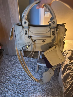 Beige Balenciaga liknande  handväska - Cool beige handväska som liknar balenciaga med detaljer som silverfärgade nitar, dragkedja framtill och avtagbar axelrem. Väskan har ett litet fack med dragkedja och en hängande kort hållare utan spegel. Perfekt för dig som gillar trendiga accessoarer.