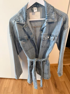 Ljusblå jeansjacka med knytskärp - Trendig ljusblå jeansjacka från H&M med knytskärp i midjan. Jackan har fyra fickor framtill, tryckknappar och klassisk krage. Perfekt oversized look och passar till många olika stilar.