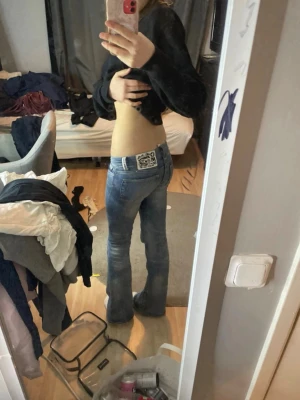 Blå bootcut jeans från weekday - Säljer ett par blå bootcut jeans från weekday med låg midja och klassisk femficksdesign. Jeansen har en snygg tvättad look och ett stort vitt logomärke bak i linningen. Perfekta för dig som gillar retrovibbar och en avslappnad stil. innerbens mått är 76 o midjan rack över är 35