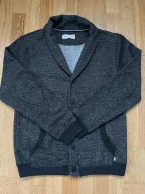 Mörkgrå kofta från Pier One - Mörkgrå kofta från Pier One med knappar framtill och snygg krage. Materialet är mjukt och har en melerad look. Koftan har förstärkta armbågslappar och två fickor. Perfekt för lager-på-lager och en stilren look.