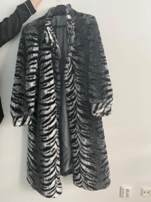 Zebramönstrad lång pälsjacka - Snygg lång pälsjacka med zebramönster i svart, vitt och grått. Jackan har en rak passform, bred krage och vida ärmar. Tillverkad i mjuk fuskpäls med slätt innerfoder. Perfekt statement-plagg för kalla vinterdagar.