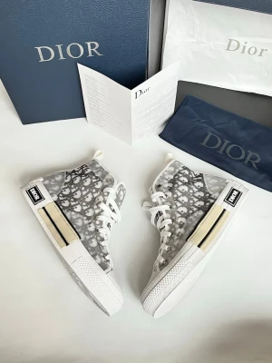 Dior B23 grå monogram sneakers - Snygga Dior B23 high-top sneakers i grått och vitt med ikoniskt Dior-monogram över hela skon. Ovandel i genomskinligt textilmaterial, vita snören och robust vit sula med svart Dior-logga på sidan. Perfekt för dig som vill sticka ut med lyxig streetstyle.