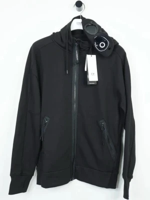 Svart hoodie från C.P. Company - Svart hoodie med dragkedja från C.P. Company. Tillverkad i mjukt material och har en modern, avslappnad passform. Perfekt för en cool streetwear-look. Helt ny med tags. 