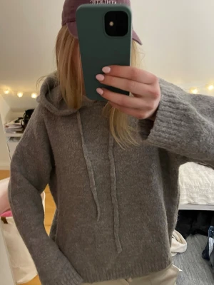 Grå stickad hoodie med huva - Mysig grå stickad hoodie med huva och dragsko. Tröjan har långa ärmar och breda ribbade muddar vid ärmslut och nederkant. Perfekt för chill dagar och enkel att matcha med jeans eller mjukisbyxor.